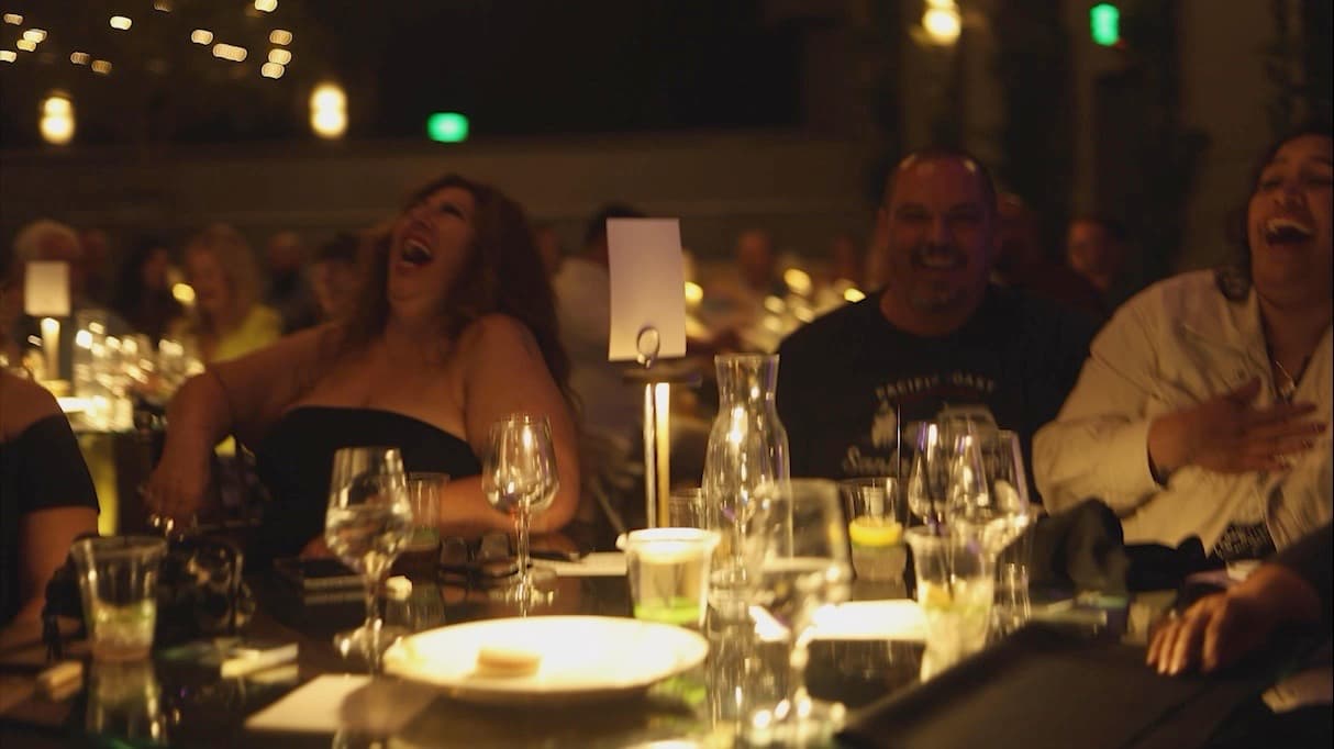 A candlelit room of people laughing at their tables during a live stand-up comedy show