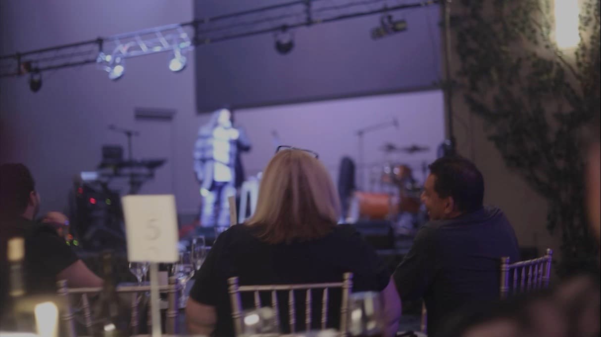 Guests at dinner tables watching a comedian on a lit stage in an event space